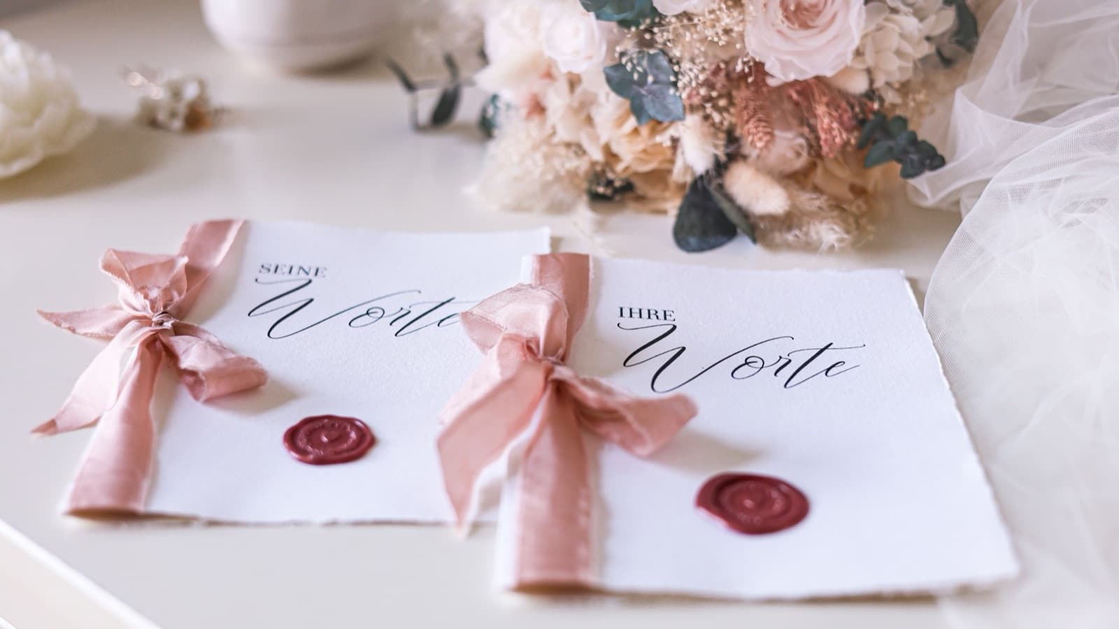 Two calligraphy cards tied with pink ribbon and sealed with red wax beside a dried flower bouquet